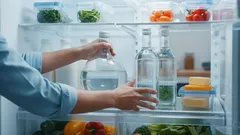 Mani che prendono bottiglia d'acqua dal frigorifero pieno di verdure e contenitori